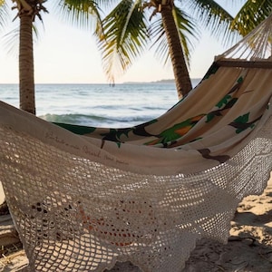 May include: A cream-colored hammock featuring a printed design of green leaves and orange accents, with intricate white crocheted fringe along the edges and wooden spreader bars, suspended between two palm trees on a sandy beach.