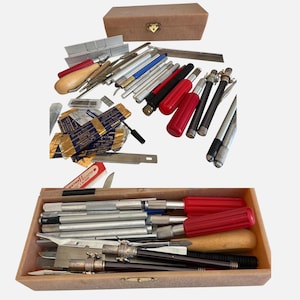 Op de afbeelding: Een verzameling van diverse hobbymessen, metalen mesjes en precisiegereedschap, sommige met rode of zwarte handvatten, naast een houten opbergdoos met een goudkleurige sluiting. De set bevat diverse losse mesjes, metalen onderdelen en een houten handvat.