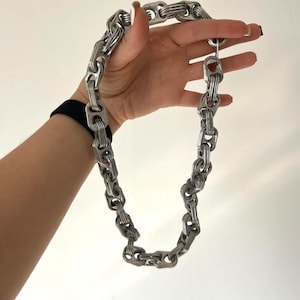 Op de afbeelding: Een zilverkleurige ketting gemaakt van gerecyclede blikjeslipjes. De schakels zijn in elkaar verweven, wat een stoere, industriële uitstraling creëert. De ketting wordt tegen een witte achtergrond gehouden. Een uniek, milieuvriendelijk accessoire.