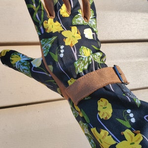 May include: A black gardening glove with a floral print featuring yellow flowers and green leaves. The glove has a brown leather cuff with a buckle.