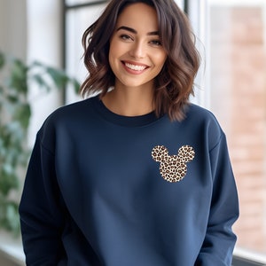 May include: Navy blue sweatshirt featuring a leopard print Mickey Mouse design on the chest. The sweatshirt has a crew neck and long sleeves. The Mickey Mouse design is made up of brown and tan spots. The background is a solid navy blue.