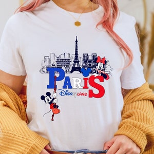 Pu&ograve; includere: Maglietta bianca con una grafica di Topolino e Minnie Mouse di fronte alla Torre Eiffel e il testo "Paris Disneyland".