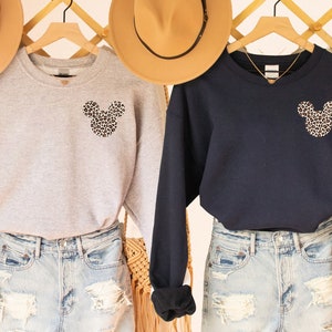 May include: Two sweatshirts, one gray and one navy blue, are displayed. Each features a leopard print Mickey Mouse head on the chest. The sweatshirts are paired with denim shorts. Hats hang in the background.