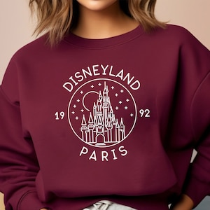 Puede incluir: Sudadera color burdeos con un gr&aacute;fico blanco del castillo de Disneyland Par&iacute;s, rodeado de estrellas y el texto "Disneyland Par&iacute;s 1992".