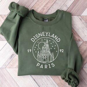 Puede incluir: Una sudadera verde con un gr&aacute;fico blanco del castillo de Disneyland Par&iacute;s, rodeado de estrellas y el texto "DISNEYLAND PARIS 1992".