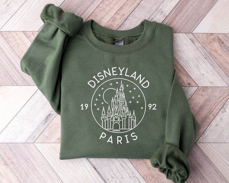 Puede incluir: Una sudadera verde con un gr&aacute;fico blanco del castillo de Disneyland Par&iacute;s, rodeado de estrellas y el texto "DISNEYLAND PARIS 1992".