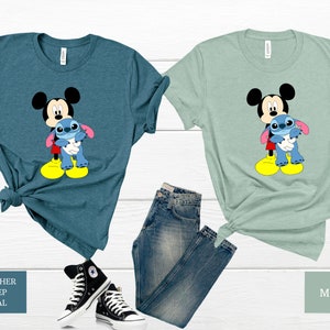 May include: Two t-shirts featuring a cartoon image of Mickey Mouse holding Stitch from Lilo & Stitch. The shirt on the left is a heather deep teal colour and the shirt on the right is a mint green colour.