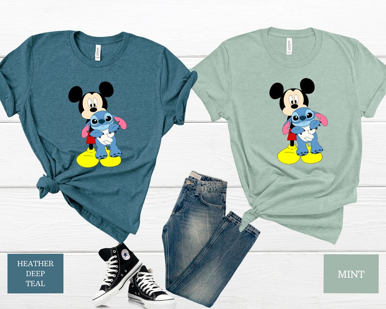 May include: Two t-shirts featuring a cartoon image of Mickey Mouse holding Stitch from Lilo & Stitch. The shirt on the left is a heather deep teal colour and the shirt on the right is a mint green colour.