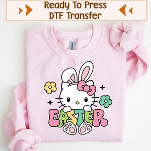 May include: A light pink sweatshirt featuring a cartoon character with bunny ears and a pink bow. The word "EASTER" is written in colorful letters below the character. The top of the image has the text "Ready To Press DTF Transfer".