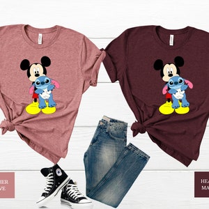 May include: Two t-shirts featuring a cartoon image of Mickey Mouse holding Stitch. The shirt on the left is heather mauve and the shirt on the right is heather maroon.