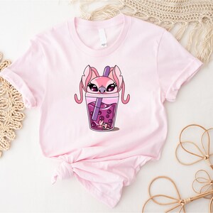 May include: A light pink t-shirt featuring a cartoon graphic of a pink character with large ears and a purple drink with boba pearls. The character is inside a cup of the drink. The shirt is made of soft material.