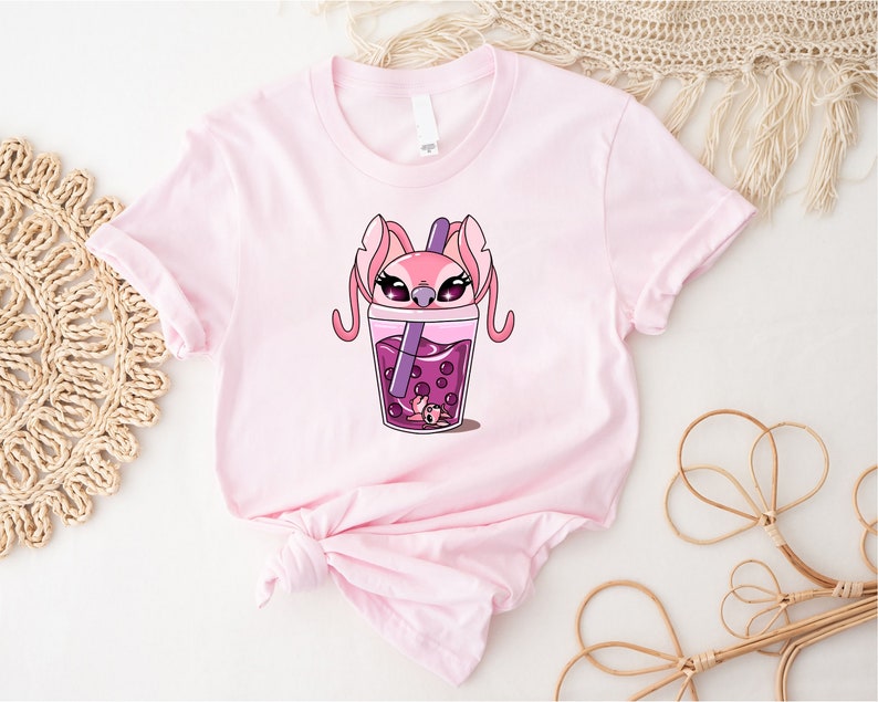 May include: A light pink t-shirt featuring a cartoon graphic of a pink character with large ears and a purple drink with boba pearls. The character is inside a cup of the drink. The shirt is made of soft material.