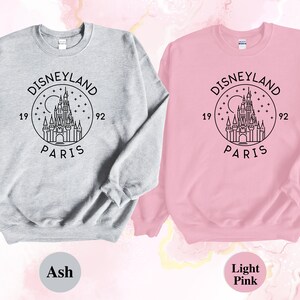 Puede incluir: Dos sudaderas, una gris ceniza y otra rosa claro, con un contorno negro del castillo de Disneyland Par&iacute;s y el texto "Disneyland Par&iacute;s 1992" en un c&iacute;rculo.