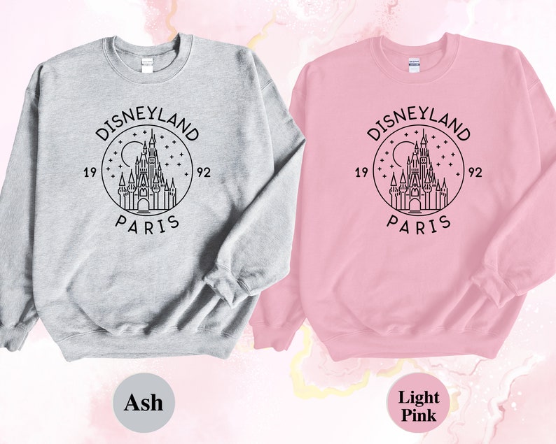 Puede incluir: Dos sudaderas, una gris ceniza y otra rosa claro, con un contorno negro del castillo de Disneyland Par&iacute;s y el texto "Disneyland Par&iacute;s 1992" en un c&iacute;rculo.