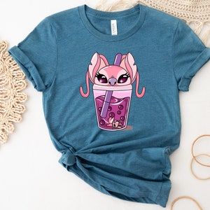 May include: Teal-colored t-shirt featuring a cartoon illustration of a pink alien character inside a boba tea cup. The boba tea is pink with dark pink boba pearls and a purple straw. The character has large eyes and long ears.