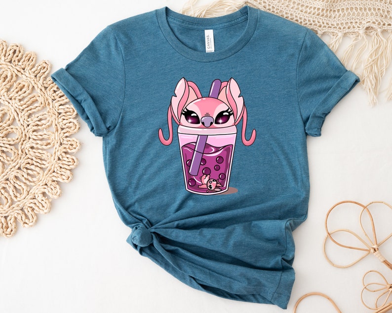 May include: Teal-colored t-shirt featuring a cartoon illustration of a pink alien character inside a boba tea cup. The boba tea is pink with dark pink boba pearls and a purple straw. The character has large eyes and long ears.