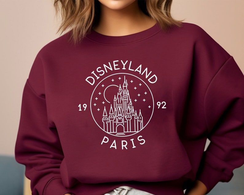 Puede incluir: Sudadera color burdeos con un gr&aacute;fico blanco del castillo de Disneyland Par&iacute;s, rodeado de estrellas y el texto "Disneyland Par&iacute;s 1992".