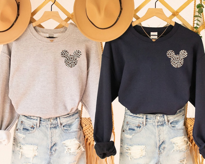 May include: Two sweatshirts, one gray and one navy blue, are displayed. Each features a leopard print Mickey Mouse head on the chest. The sweatshirts are paired with denim shorts. Hats hang in the background.