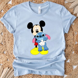 May include: Light blue t-shirt featuring a cartoon image of Mickey Mouse holding Stitch from Lilo & Stitch.