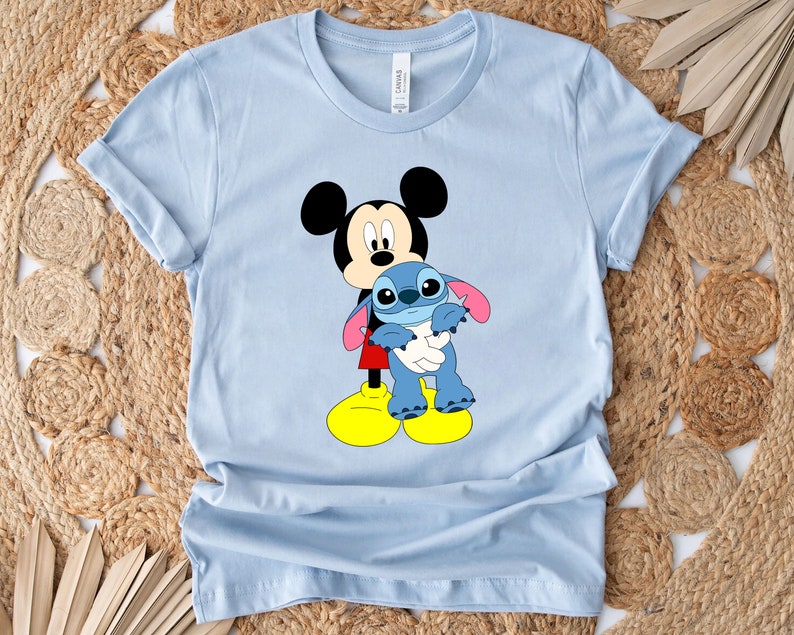 May include: Light blue t-shirt featuring a cartoon image of Mickey Mouse holding Stitch from Lilo & Stitch.