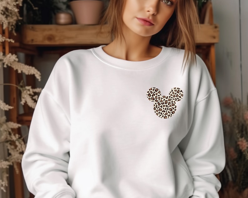 May include: White sweatshirt featuring a leopard print Mickey Mouse head design on the chest. The design has a brown and tan spotted pattern. The sweatshirt has long sleeves and a crew neck.