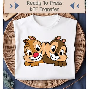 May include: White t-shirt with a cartoon graphic of Chip and Dale, the Disney chipmunks. The image shows the chipmunks smiling. The text "Ready To Press DTF Transfer" is at the top. The shirt is on a woven basket.