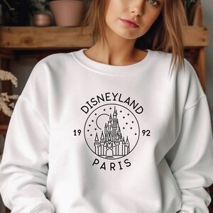 Puede incluir: Sudadera blanca con un gr&aacute;fico negro del logotipo de Disneyland Par&iacute;s, que presenta un dibujo estilizado del castillo de la Bella Durmiente, el texto "DISNEYLAND PARIS" y los a&ntilde;os "1992" y "19".