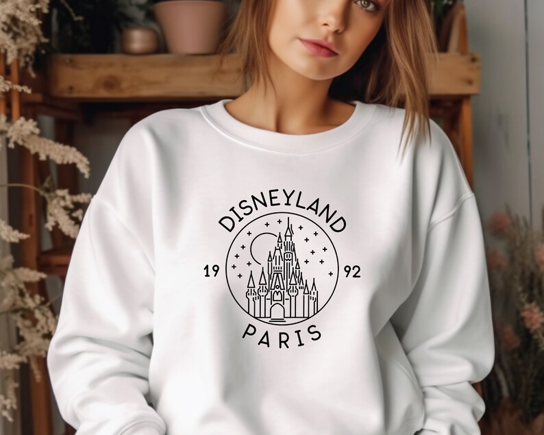 Puede incluir: Sudadera blanca con un gr&aacute;fico negro del logotipo de Disneyland Par&iacute;s, que presenta un dibujo estilizado del castillo de la Bella Durmiente, el texto "DISNEYLAND PARIS" y los a&ntilde;os "1992" y "19".