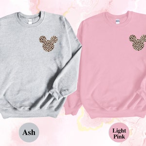 May include: Two sweatshirts, one ash gray and one light pink, each featuring a leopard print Mickey Mouse head design on the upper left chest. The sweatshirts are displayed against a soft, abstract background.
