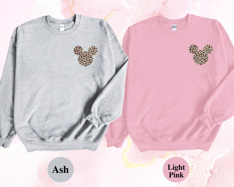 May include: Two sweatshirts, one ash gray and one light pink, each featuring a leopard print Mickey Mouse head design on the upper left chest. The sweatshirts are displayed against a soft, abstract background.