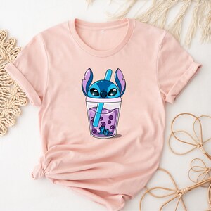 May include: A light pink t-shirt featuring a cartoon graphic of a blue alien character in a boba drink. The drink is purple with dark purple boba pearls and a blue straw. The character's head and ears are visible above the cup.