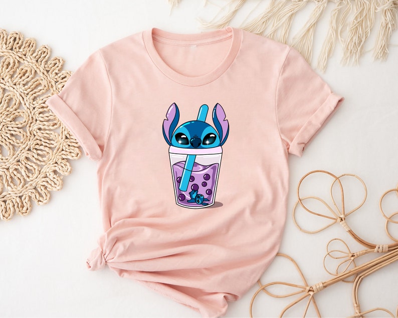 May include: A light pink t-shirt featuring a cartoon graphic of a blue alien character in a boba drink. The drink is purple with dark purple boba pearls and a blue straw. The character's head and ears are visible above the cup.