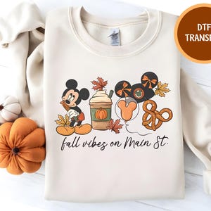 Può includere: Felpa color crema con grafica a tema autunnale con Topolino, un pumpkin spice latte, palloncini e un pretzel. Il testo "fall vibes on Main St." è scritto in corsivo. Un'etichetta di trasferimento DTF si trova nell'angolo in alto a destra.