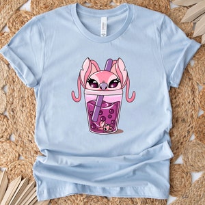 May include: Light blue t-shirt featuring a cartoon graphic of a pink alien character inside a purple boba tea drink. The character has large eyes and long ears. The drink has purple boba pearls and a purple straw.