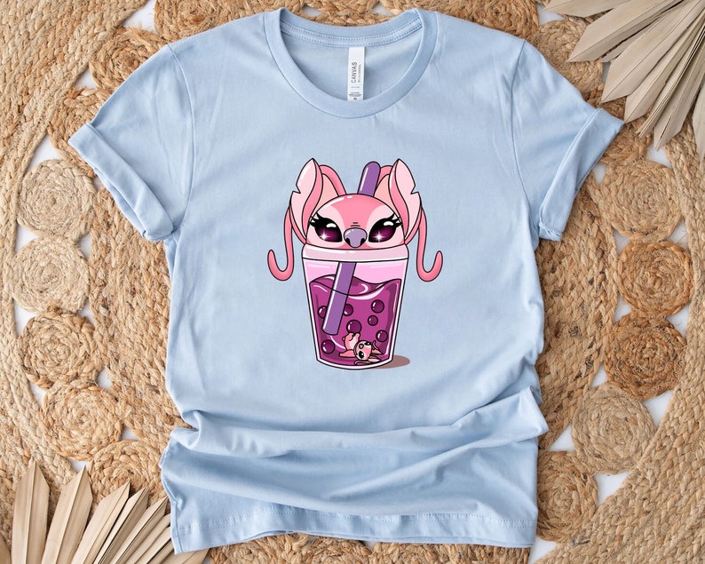 May include: Light blue t-shirt featuring a cartoon graphic of a pink alien character inside a purple boba tea drink. The character has large eyes and long ears. The drink has purple boba pearls and a purple straw.