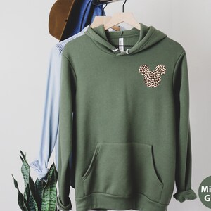 May include: Olive green hoodie with a kangaroo pocket and a leopard print Mickey Mouse head design on the chest. The hoodie is hanging on a wooden hanger with other clothing items. The sleeve cuffs are ribbed.