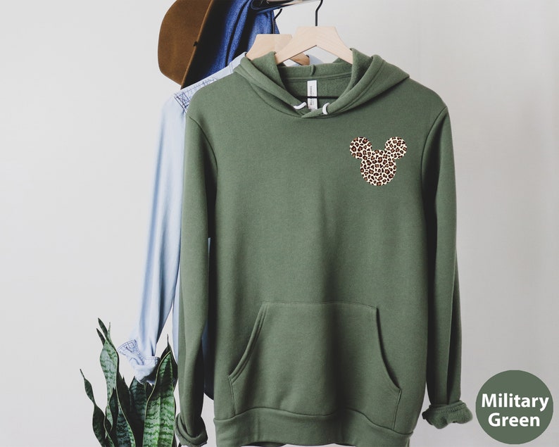 May include: Olive green hoodie with a kangaroo pocket and a leopard print Mickey Mouse head design on the chest. The hoodie is hanging on a wooden hanger with other clothing items. The sleeve cuffs are ribbed.