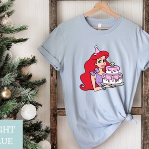 Pode incluir: Camiseta azul claro com um gr&aacute;fico de um personagem de desenho animado de cabelo ruivo usando um chap&eacute;u de anivers&aacute;rio e segurando um bolo rosa e branco.