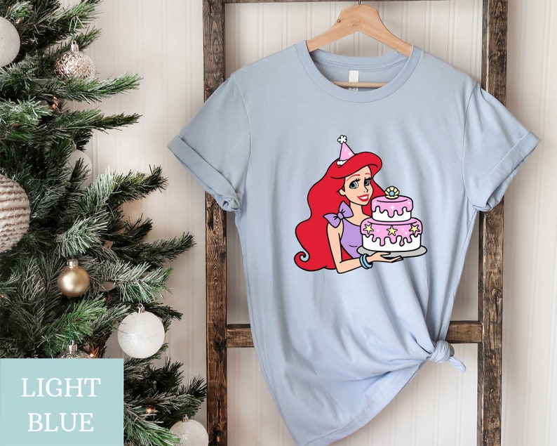 Pode incluir: Camiseta azul claro com um gr&aacute;fico de um personagem de desenho animado de cabelo ruivo usando um chap&eacute;u de anivers&aacute;rio e segurando um bolo rosa e branco.