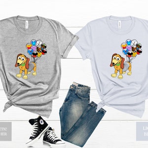 May include: A grey and a light blue t-shirt featuring a cartoon dog holding balloons. The balloons are shaped like various Disney characters. The t-shirts are shown against a white background with a pair of black trainers and blue jeans.