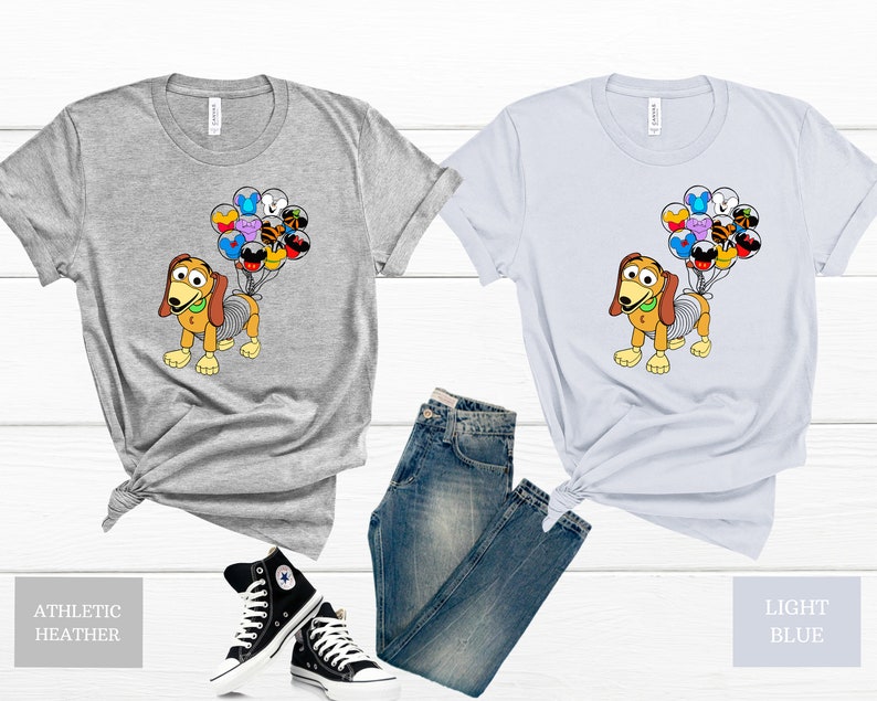 May include: A grey and a light blue t-shirt featuring a cartoon dog holding balloons. The balloons are shaped like various Disney characters. The t-shirts are shown against a white background with a pair of black trainers and blue jeans.