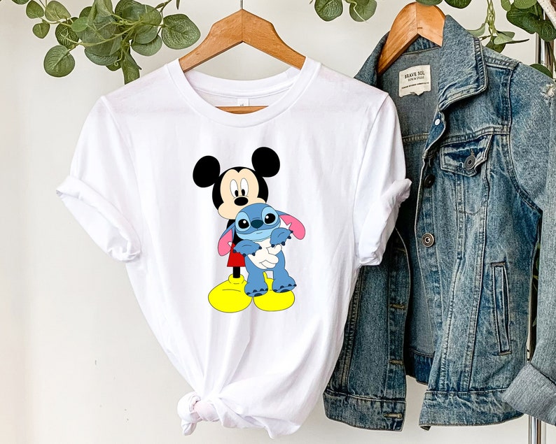 May include: White t-shirt featuring a graphic of Mickey Mouse holding Stitch, a blue alien character from the Disney film Lilo & Stitch.