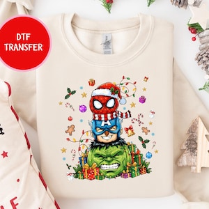 Pode incluir: Camisola creme com uma transferência DTF de Homem-Aranha, Capitão América e Hulk empilhados, todos usando chapéus de Natal. O design inclui luzes de Natal, enfeites e presentes.