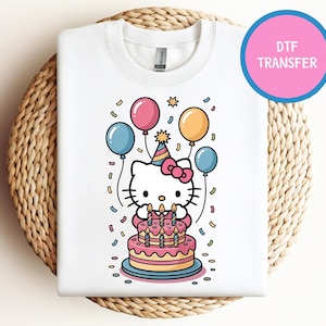 May include: White sweatshirt featuring a colorful graphic of a cartoon cat celebrating a birthday. The cat is wearing a party hat and surrounded by balloons and confetti. A pink cake with lit candles is in front of the cat. The text "DTF TRANSFER" is in a pink circle.