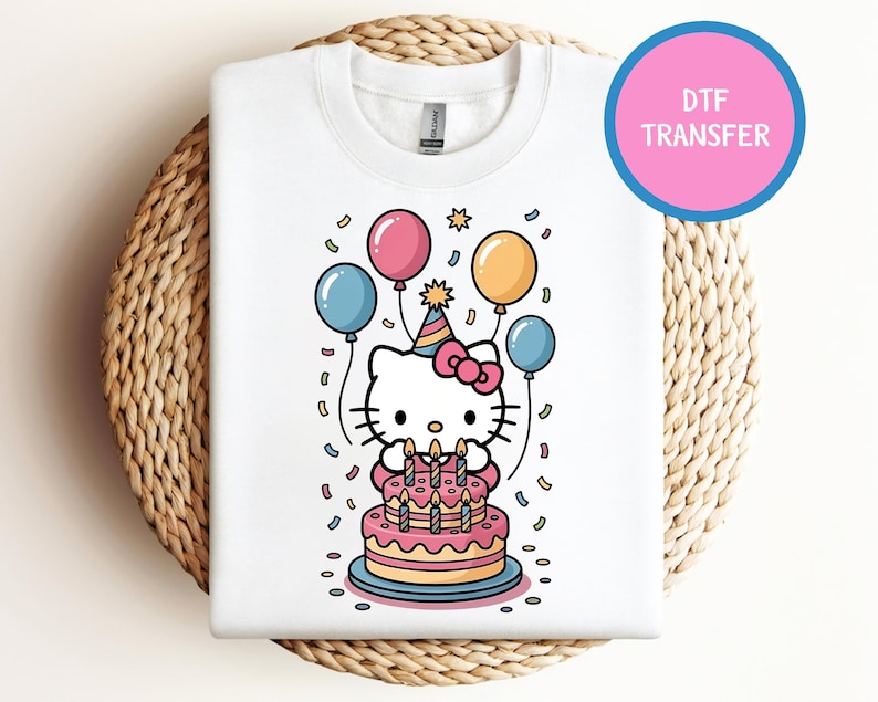 May include: White sweatshirt featuring a colorful graphic of a cartoon cat celebrating a birthday. The cat is wearing a party hat and surrounded by balloons and confetti. A pink cake with lit candles is in front of the cat. The text "DTF TRANSFER" is in a pink circle.