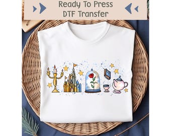 Könnte beinhalten: Weißes T-Shirt mit einem DTF-Transfer-Design, das ein gelbes Schloss, eine Rose in einer Glaskuppel, eine Teekanne, eine Tasse und einen Kerzenhalter zeigt. Das Design ist von blauen und gelben Aquarellspritzer und Sternen umgeben.
