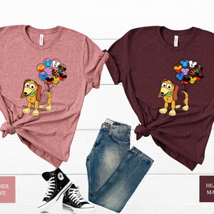 May include: Two t-shirts featuring a cartoon dog holding balloons with Disney character icons. The shirt on the left is heather mauve and the shirt on the right is heather maroon.