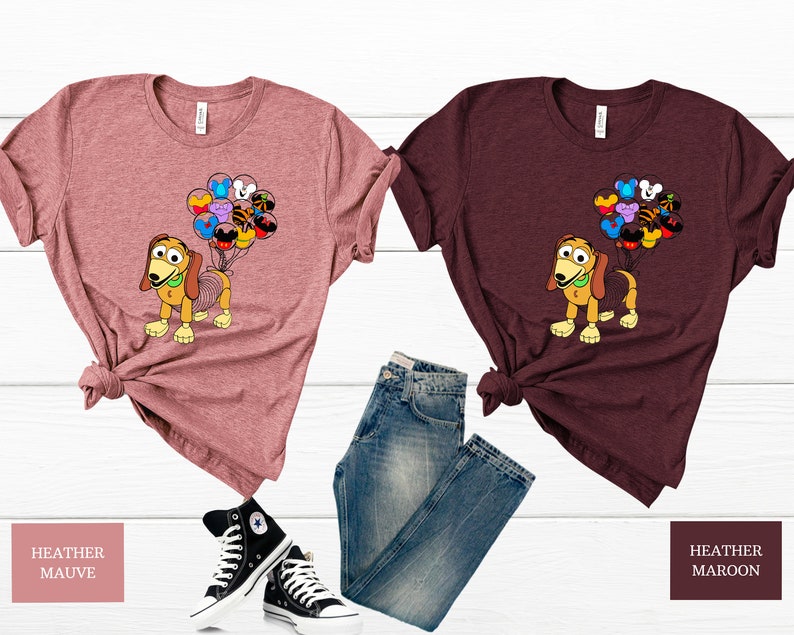 May include: Two t-shirts featuring a cartoon dog holding balloons with Disney character icons. The shirt on the left is heather mauve and the shirt on the right is heather maroon.