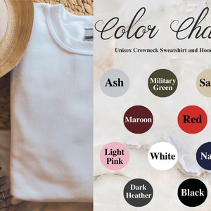 May include: A white unisex crewneck sweatshirt, folded neatly, with a straw hat and woven basket in the background. A color chart displays various sweatshirt colors: Ash, Military Green, Sand, Maroon, Red, Light Pink, White, Navy, Dark Heather, and Black.