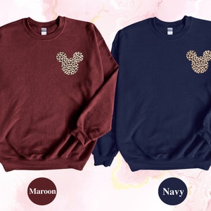 May include: Two sweatshirts, one maroon and one navy, each featuring a leopard print Mickey Mouse head design on the upper left chest. The maroon sweatshirt has the word "Maroon" below it. The navy sweatshirt has the word "Navy" below it.